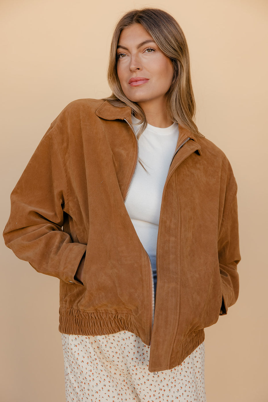 Woman wearing a brown suede jacket over a white top and patterned skirt on a beige background