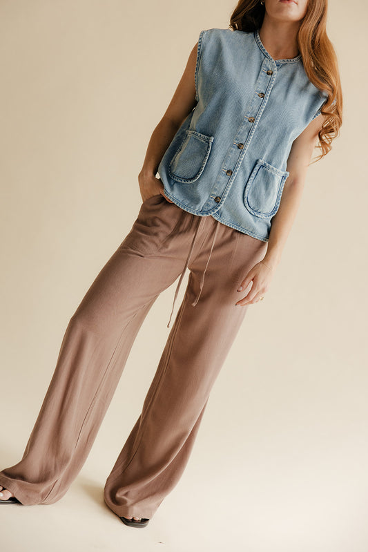 Woman wearing a denim vest over a brown top and wide-leg pants on a beige background
