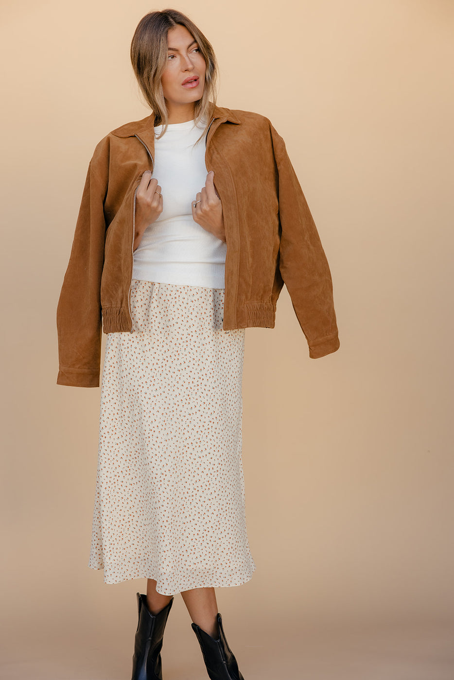 Woman wearing a brown jacket, white top, and patterned skirt against a beige background