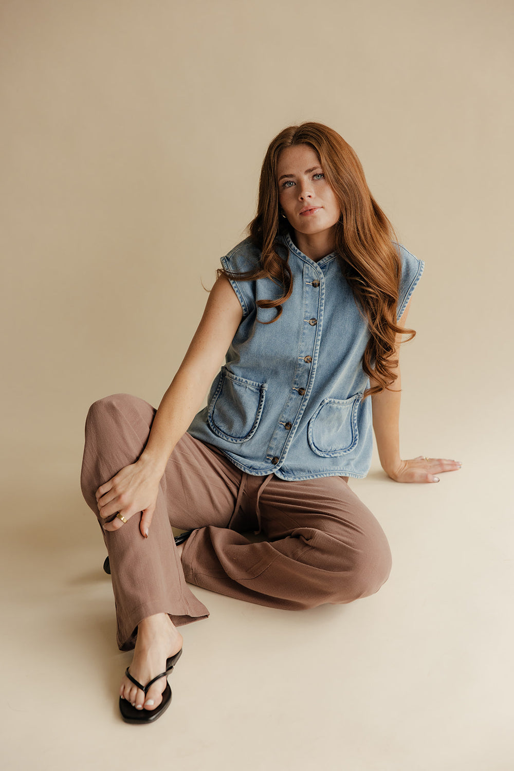 Woman wearing a denim vest and brown pants sitting on a beige floor.