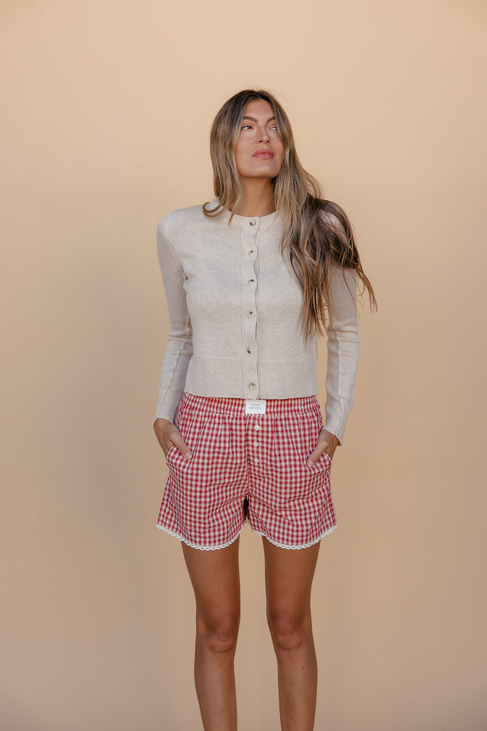 Woman wearing a beige cardigan and red checkered shorts against a beige background