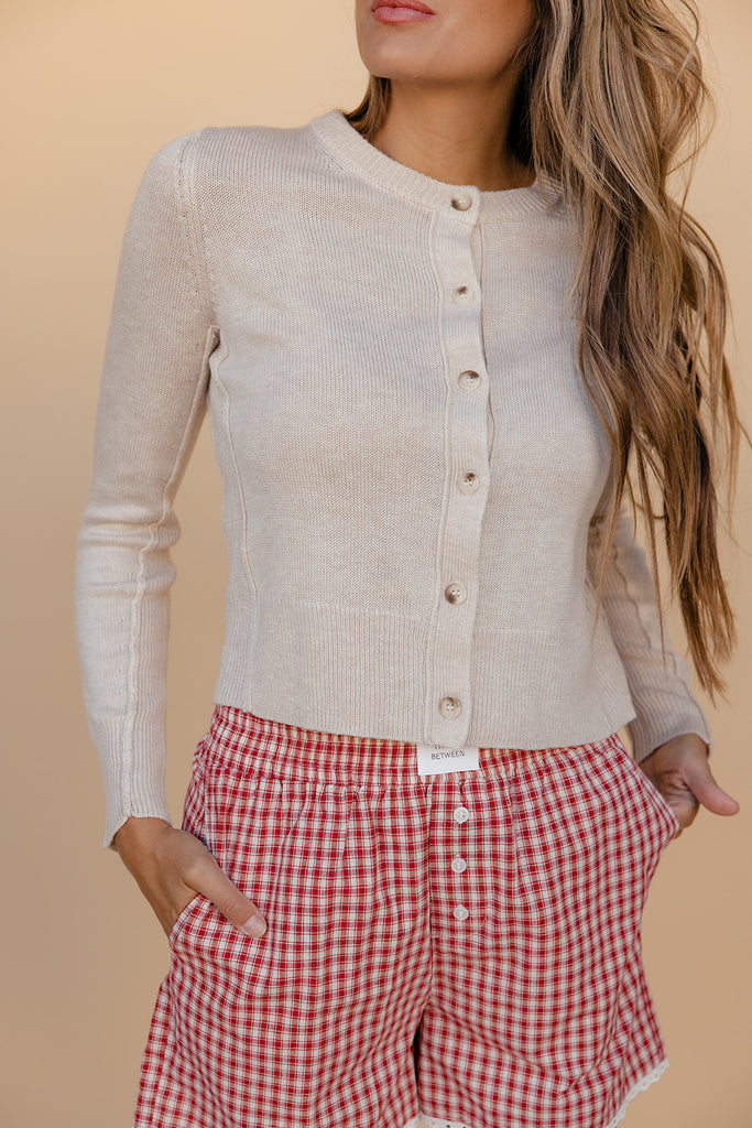 Woman wearing a beige cardigan over a white shirt with red checkered pants on a beige background