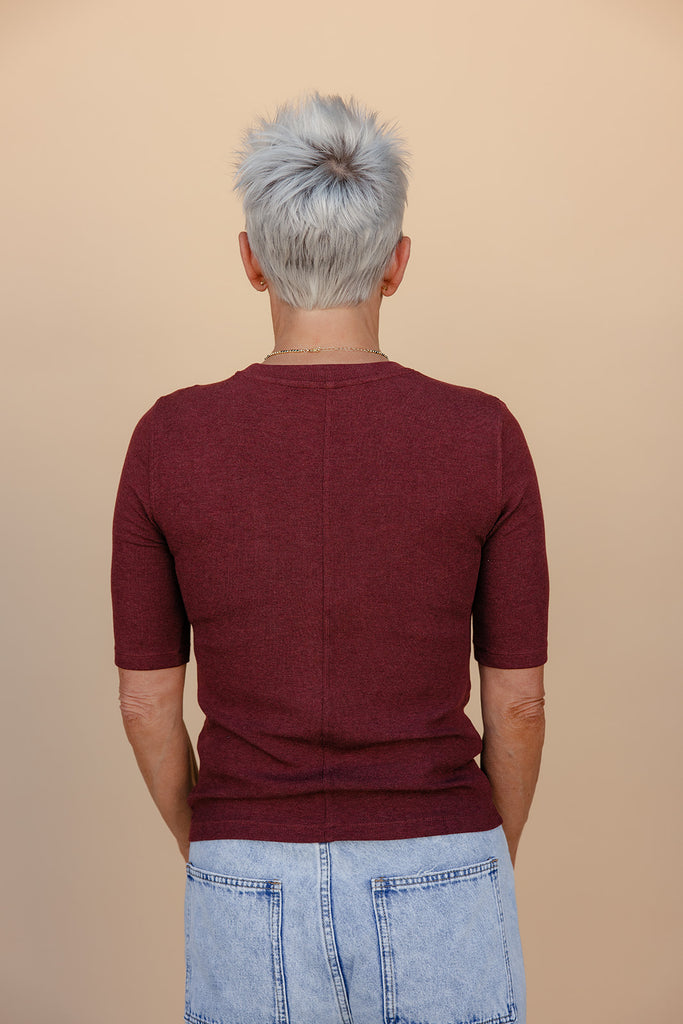 Person wearing a maroon sweater and light blue jeans with gray hair, standing against a beige background