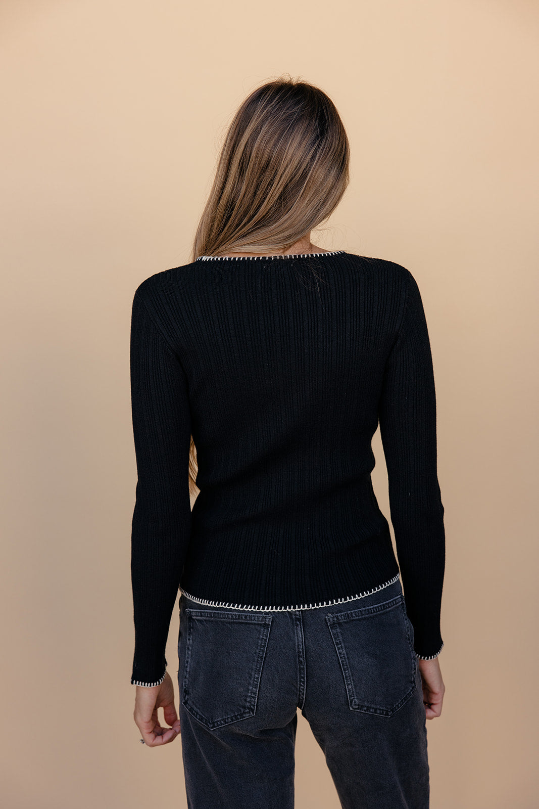 Person wearing a black long-sleeve top and dark jeans against a beige background