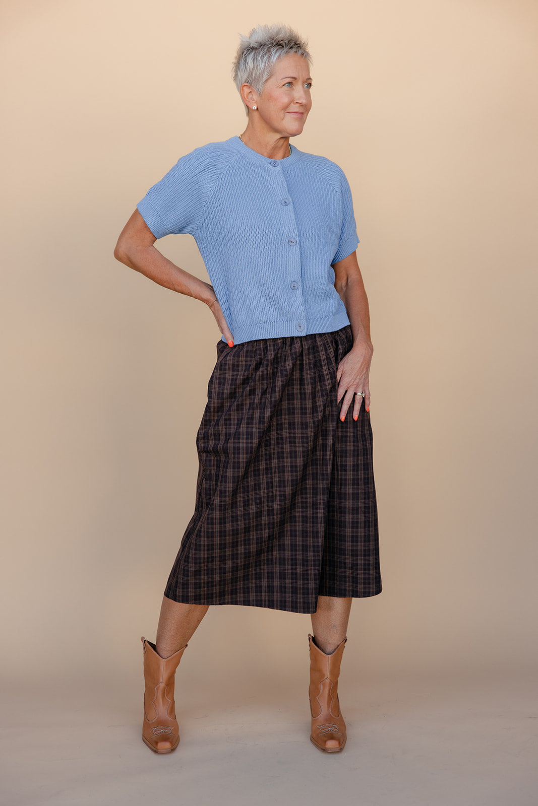 Woman wearing a blue cardigan, plaid skirt, and brown boots against a beige background