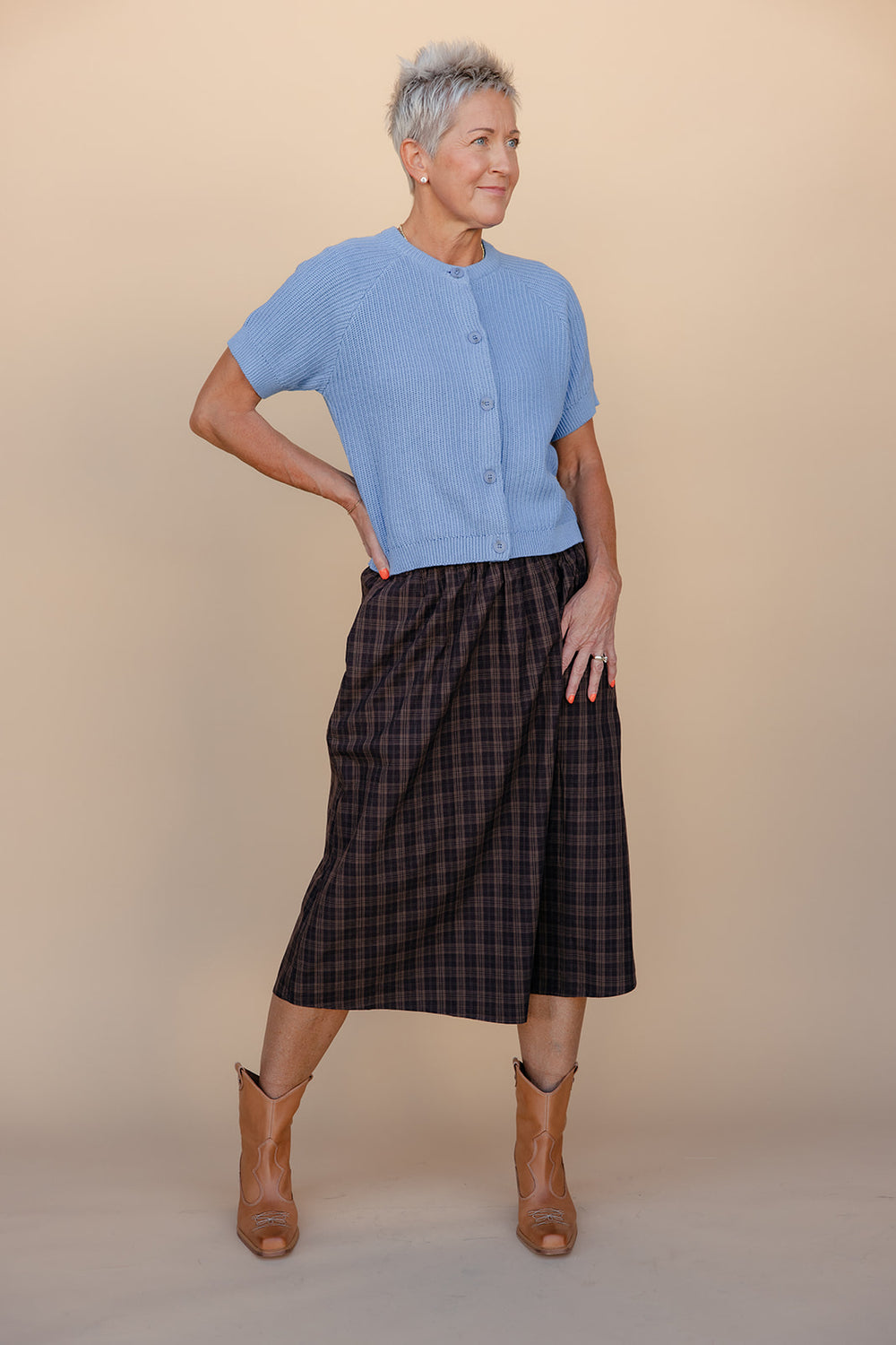 Woman wearing a blue cardigan, plaid skirt, and brown boots against a beige background