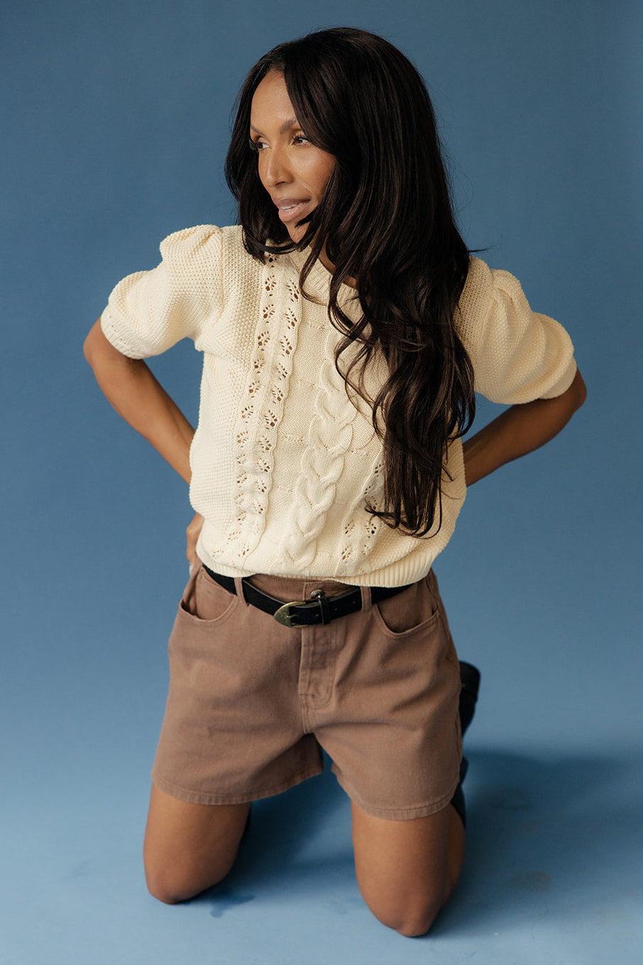 Woman kneeling on a blue background wearing a short sleeve sweater