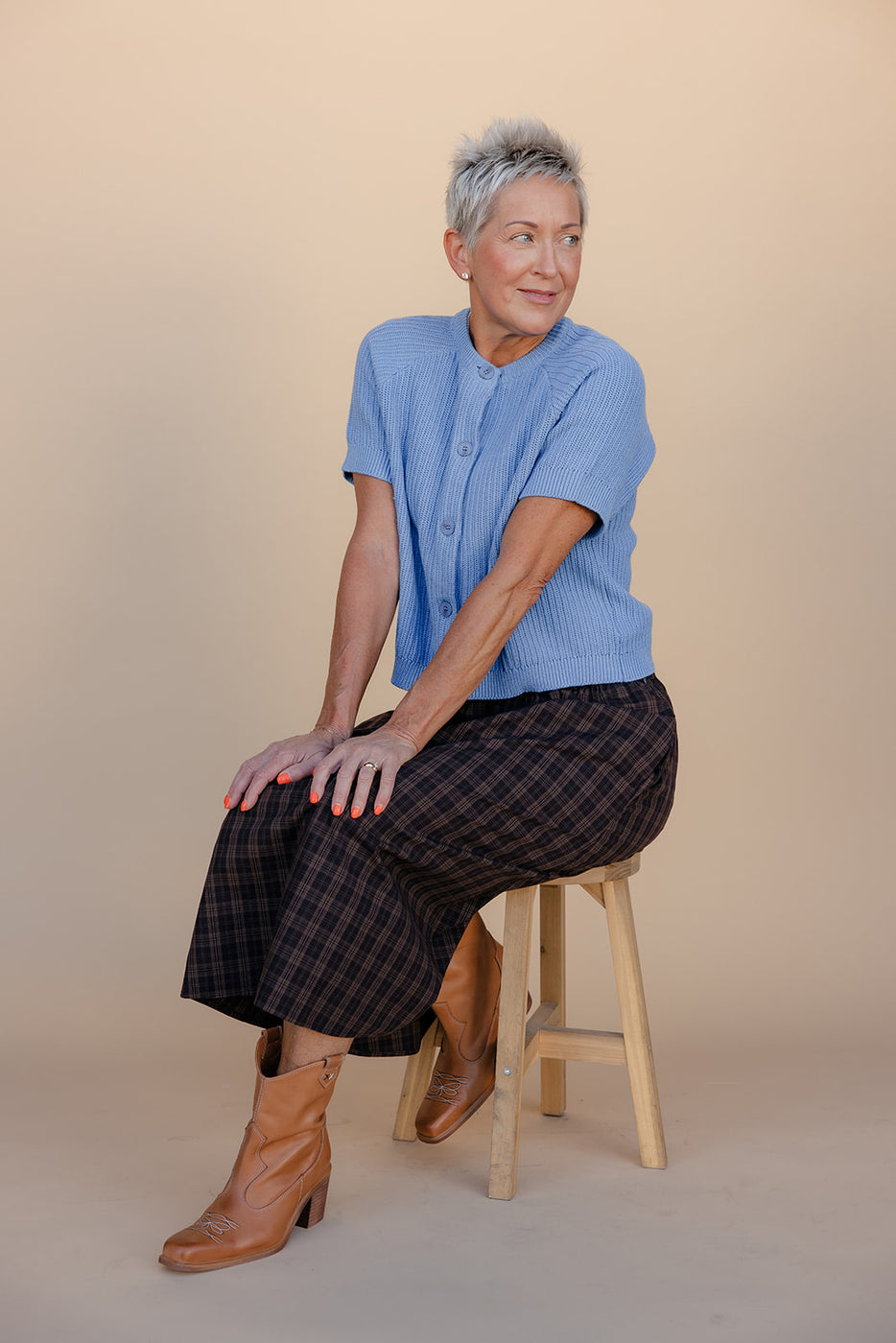 Woman sitting on a stool wearing a blue shirt and plaid skirt against a beige background