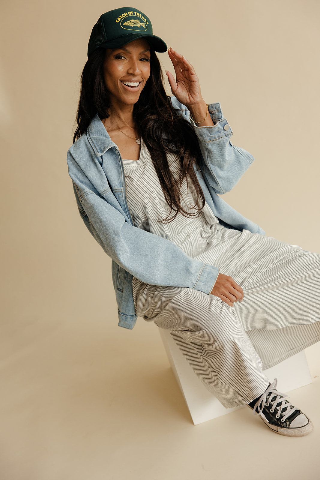 Woman wearing a denim jacket, white dress, and black sneakers on a beige background