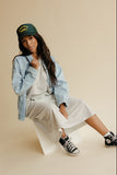 Woman sitting on a white stool wearing a denim jacket, white dress, and black sneakers on a beige background