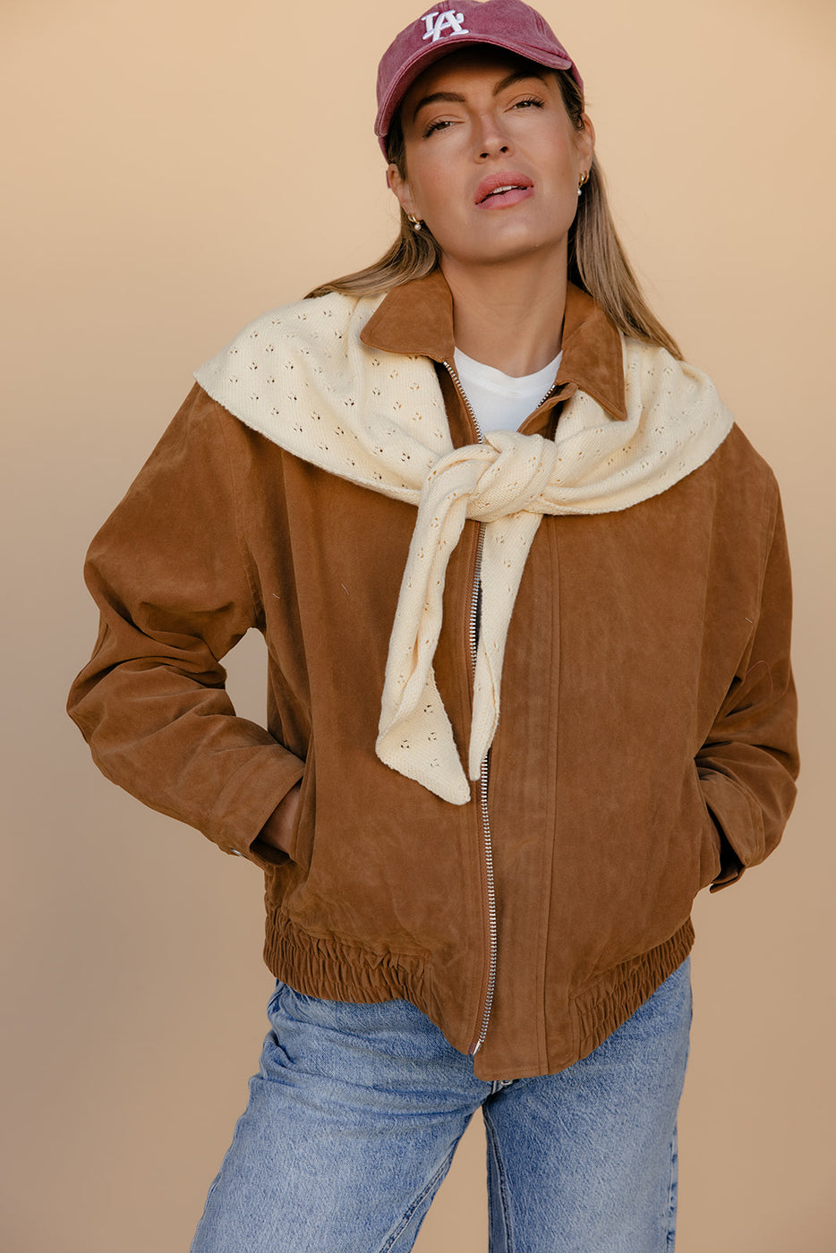 Woman wearing a brown jacket, white scarf, blue jeans, and a purple cap against a beige background