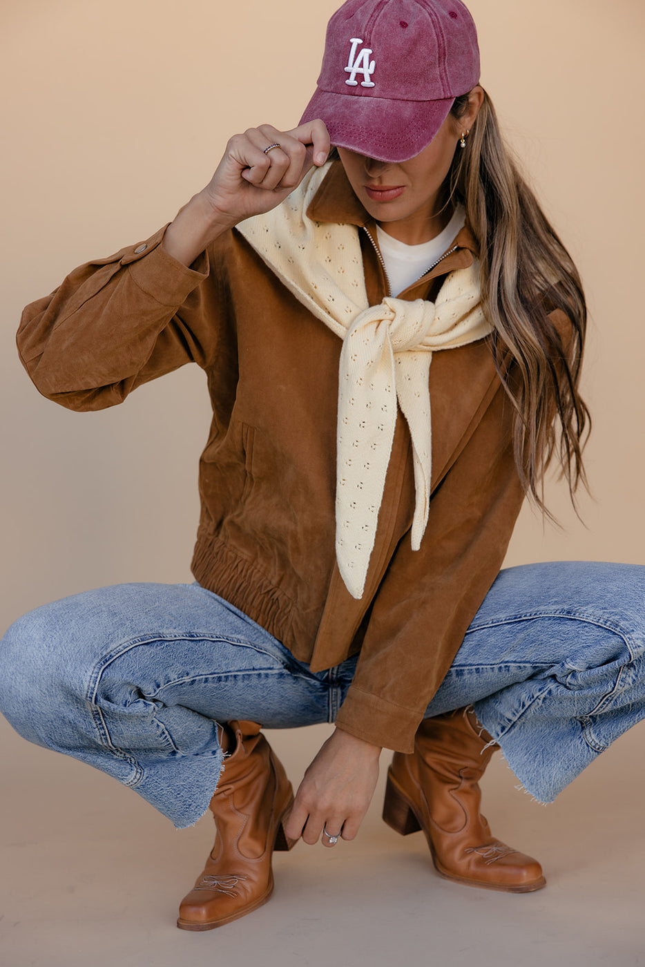 Person wearing a brown vest, blue jeans, brown boots, and a pink cap with 'LA' on a beige background