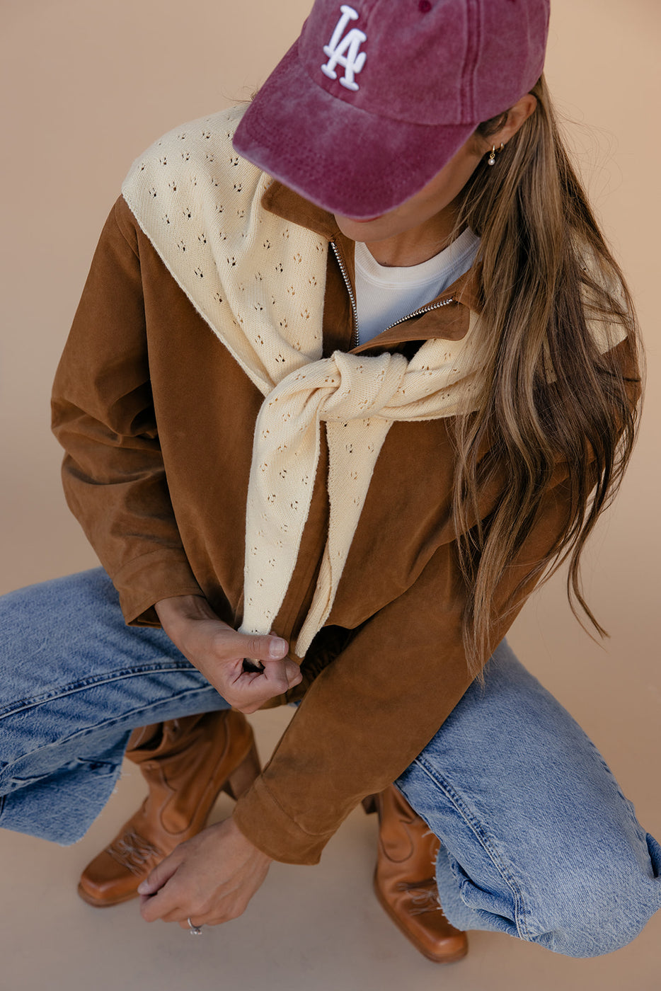 Person wearing a brown jacket, blue jeans, and a maroon cap with 'LA' on a beige background