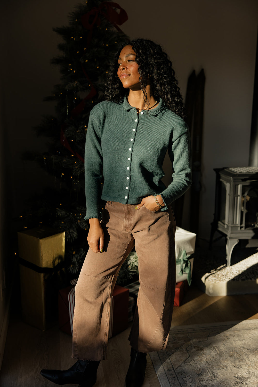 Woman in a green sweater and brown pants standing in a room with a Christmas tree.