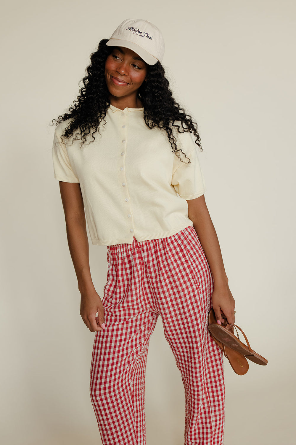 Woman wearing a white shirt, red checkered pants, and a white cap with a logo on a plain background