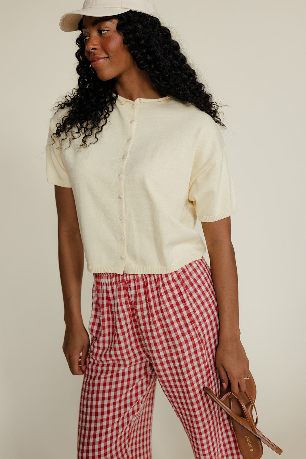 Woman wearing a white shirt and red checkered pants with a beige cap on a plain background