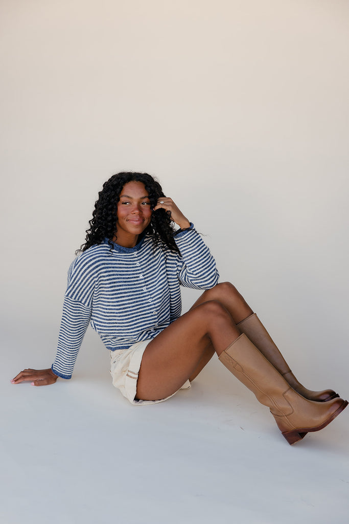 Woman sitting on a white floor wearing a striped sweater, shorts, and knee-high boots.