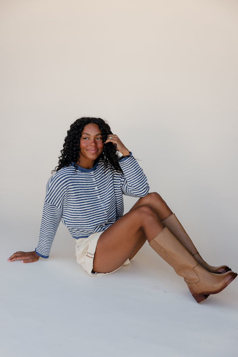 Woman sitting on a white floor wearing a striped sweater, shorts, and knee-high boots.