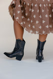 Person wearing a brown floral dress and black cowboy boots on a plain background