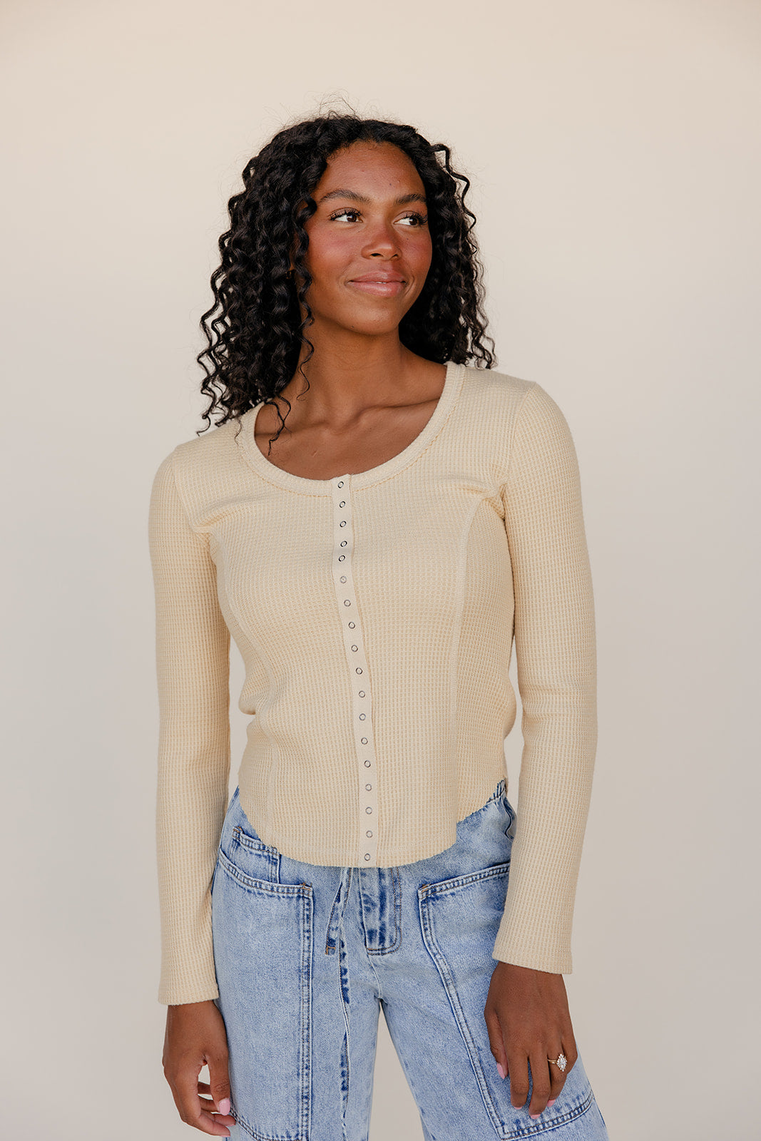 Woman wearing a beige long-sleeve top and blue jeans against a plain background