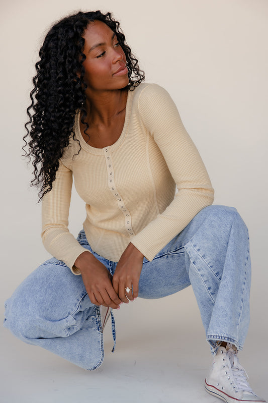 Woman wearing a beige long-sleeve top and blue jeans sitting on the floor against a plain background