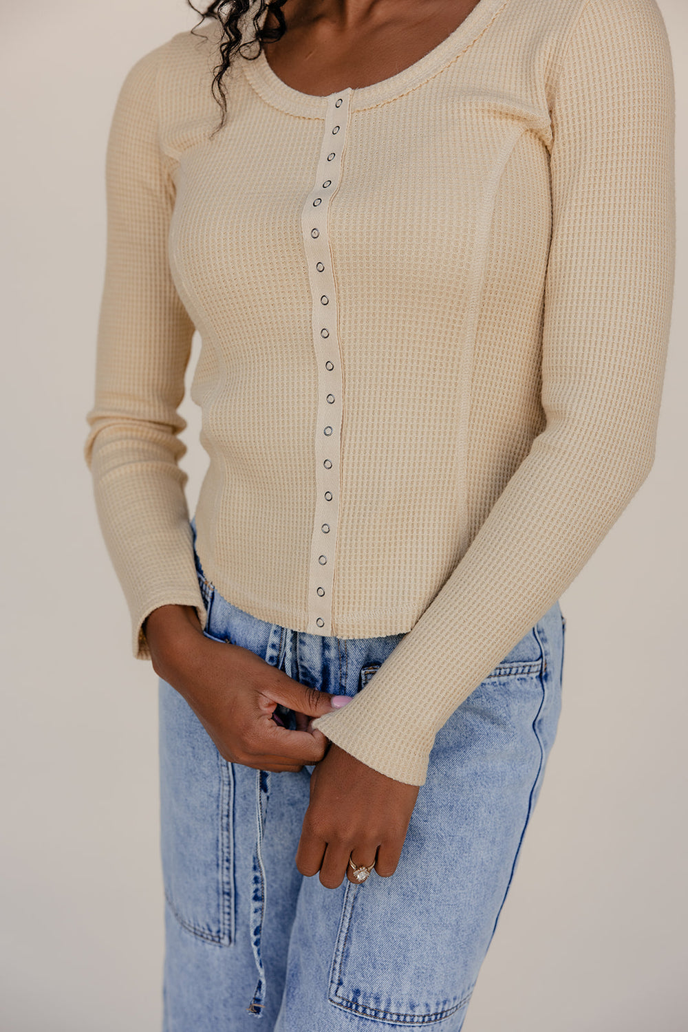Person wearing a beige long-sleeve top with button details and blue jeans on a plain background
