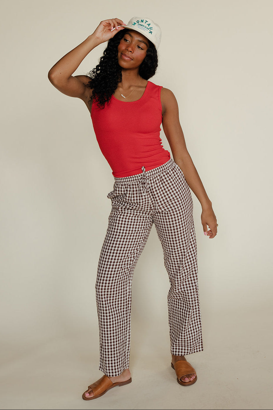 Woman wearing a red tank top and checkered pants with a white cap on a beige background
