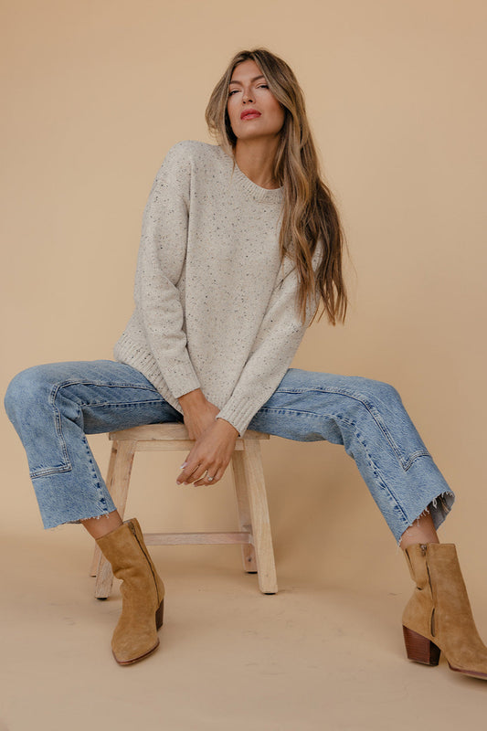 Woman sitting on a stool wearing a light gray sweater, blue jeans, and brown boots against a beige background