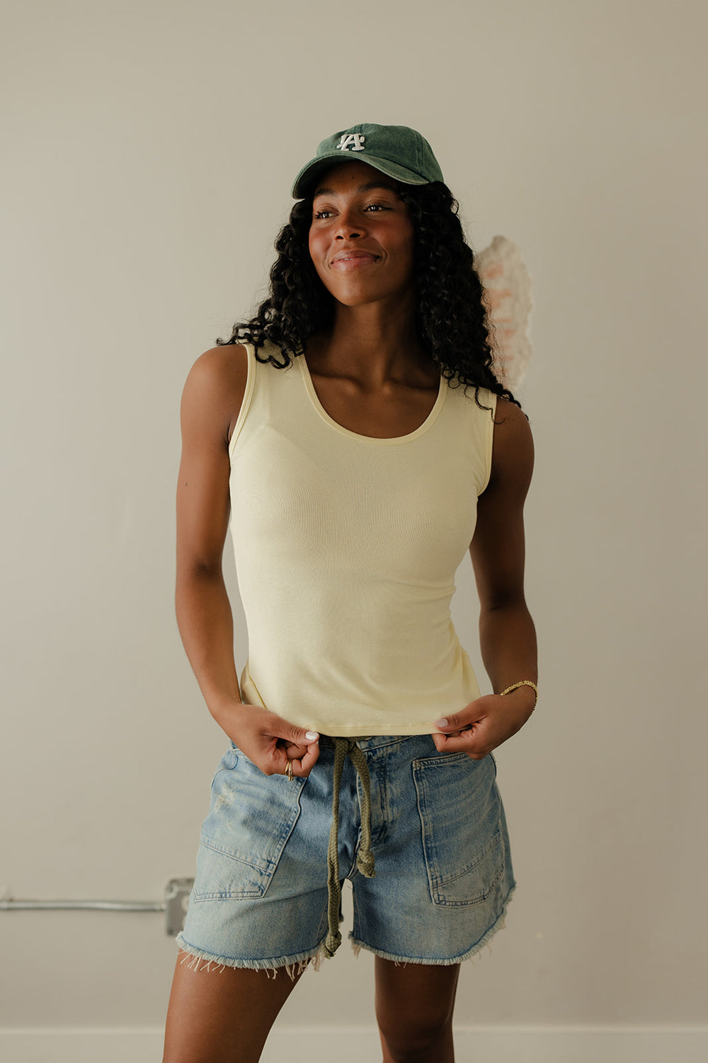 Woman wearing a beige tank top and denim shorts with a green cap indoors.