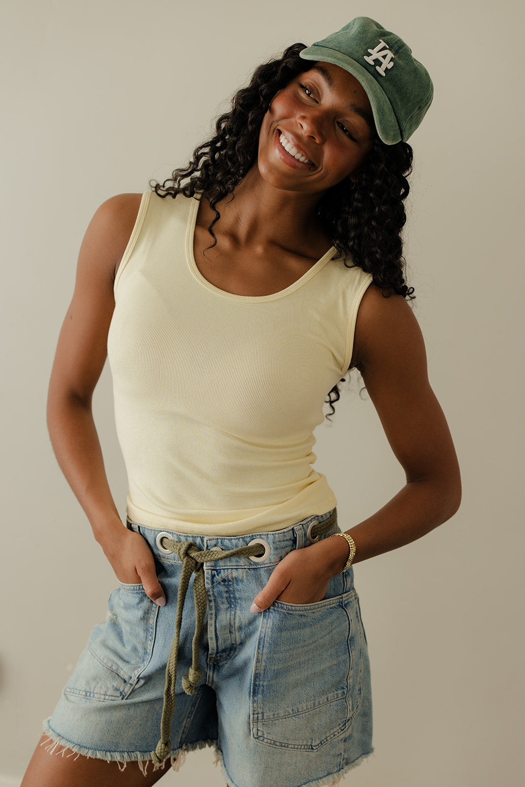 Woman wearing a yellow tank top, denim shorts, and a green cap with a logo on a plain background