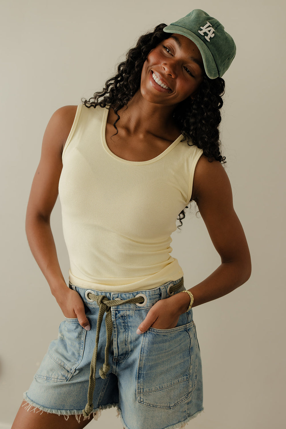 Woman wearing a yellow tank top, denim shorts, and a green cap with a logo on a plain background