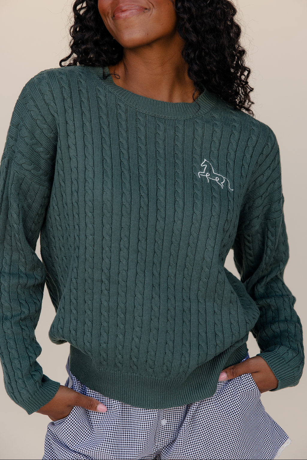 Person wearing a green cable knit sweater with a logo on a plain background