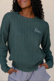 Person wearing a green cable knit sweater with a logo on a plain background