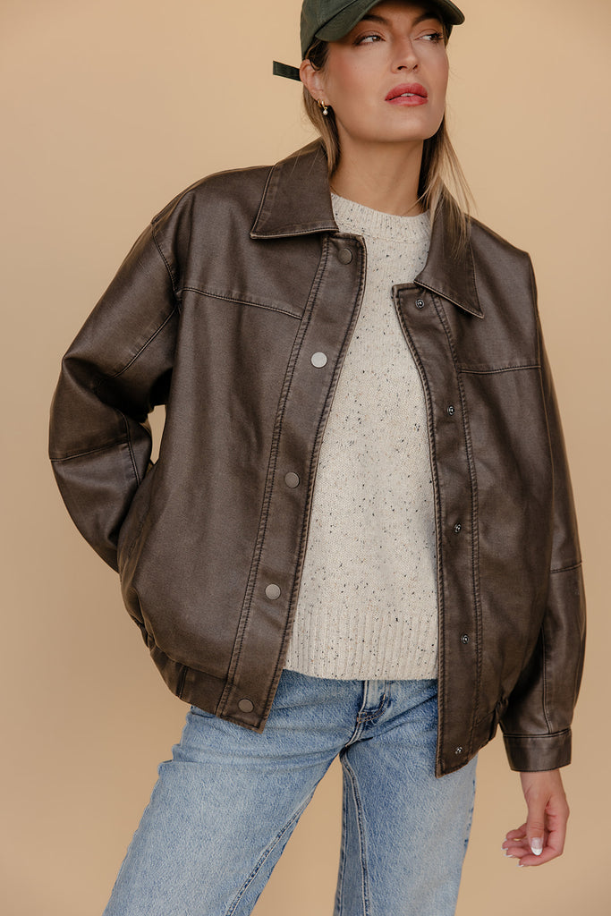 Woman wearing a brown leather jacket, white sweater, and blue jeans against a beige background
