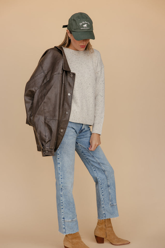 Person wearing a brown jacket, gray sweater, blue jeans, and tan boots against a beige background