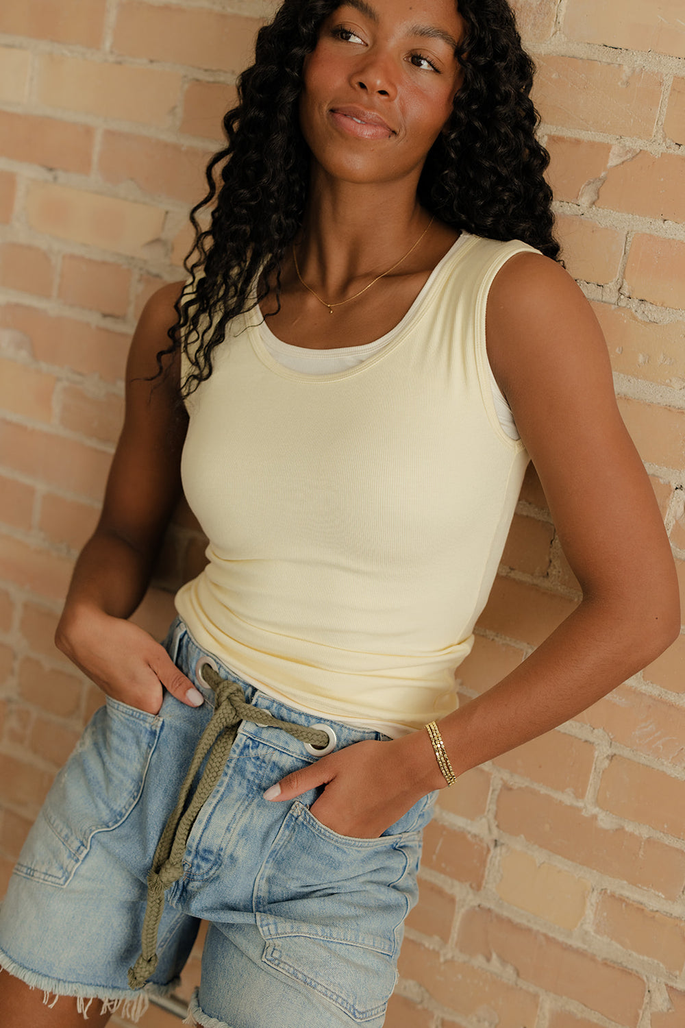 Woman wearing a beige tank top and denim shorts against a brick wall.
