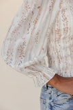 Close-up of a person wearing a light-colored blouse with floral patterns and blue jeans on a beige background.
