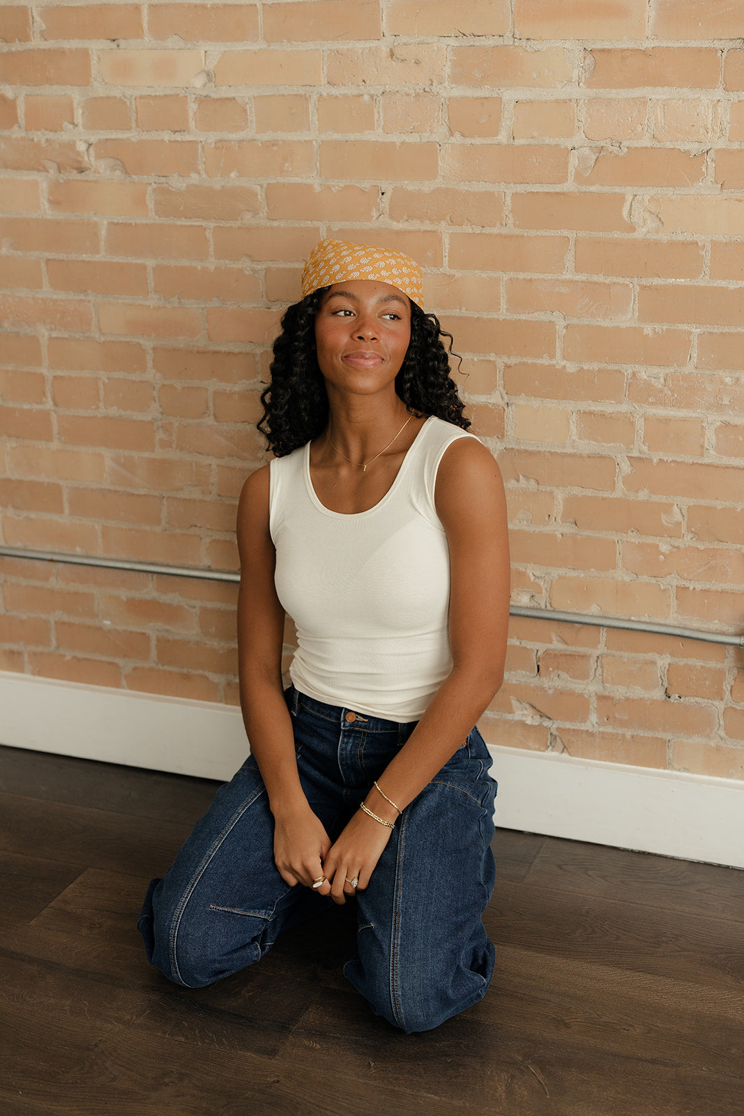 Woman kneeling in front of a brick wall wearing a white tank top and blue jeans.