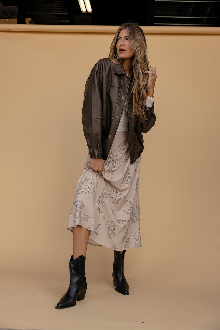 Woman wearing a dark jacket, patterned dress, and black boots against a beige background
