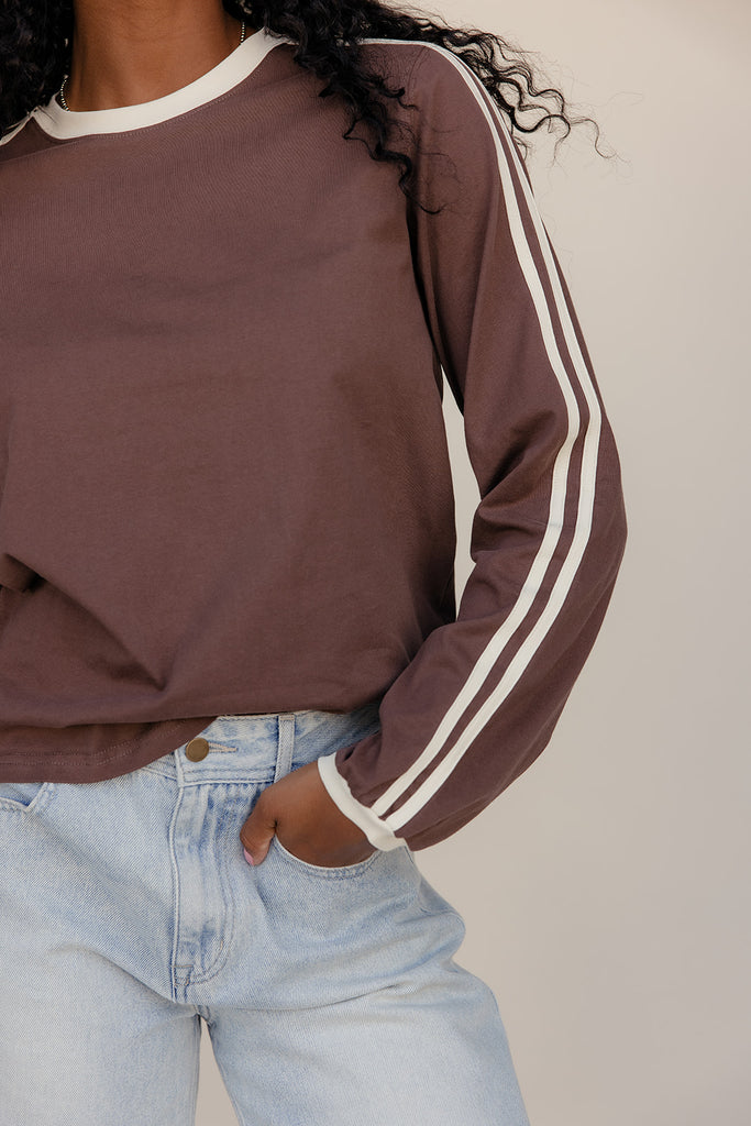 Person wearing a brown sweatshirt with white stripes on sleeves and light blue jeans against a neutral background