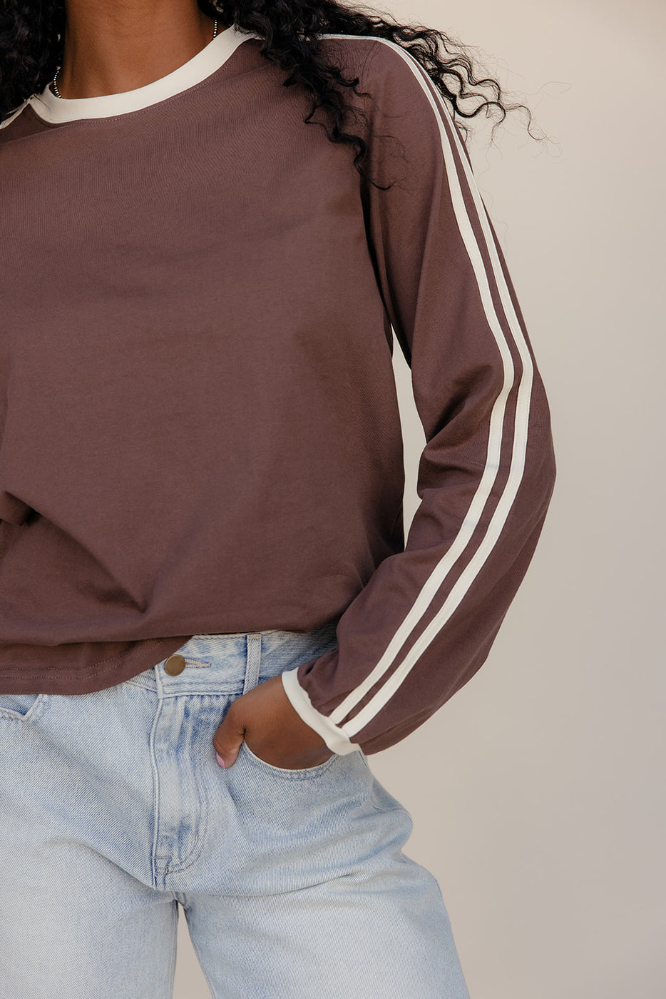 Person wearing a brown sweatshirt with white stripes on sleeves and light blue jeans against a neutral background