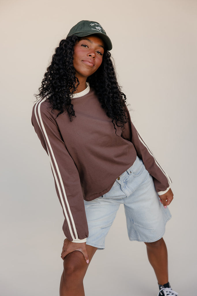 Person wearing a brown sweatshirt with white stripes and light blue shorts against a plain background