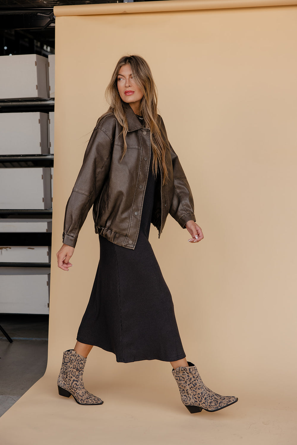 Woman wearing a brown leather jacket and black dress with patterned boots on a beige background