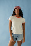 Person wearing a beige ringer t-shirt with orange trim and a red cap against a blue background