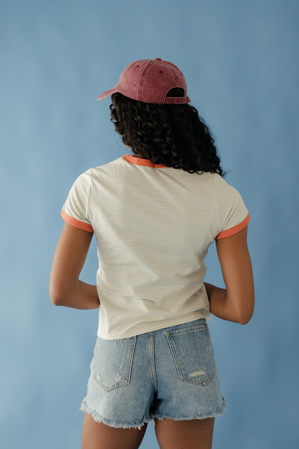 Person wearing a striped shirt and denim shorts with a maroon cap on a blue background