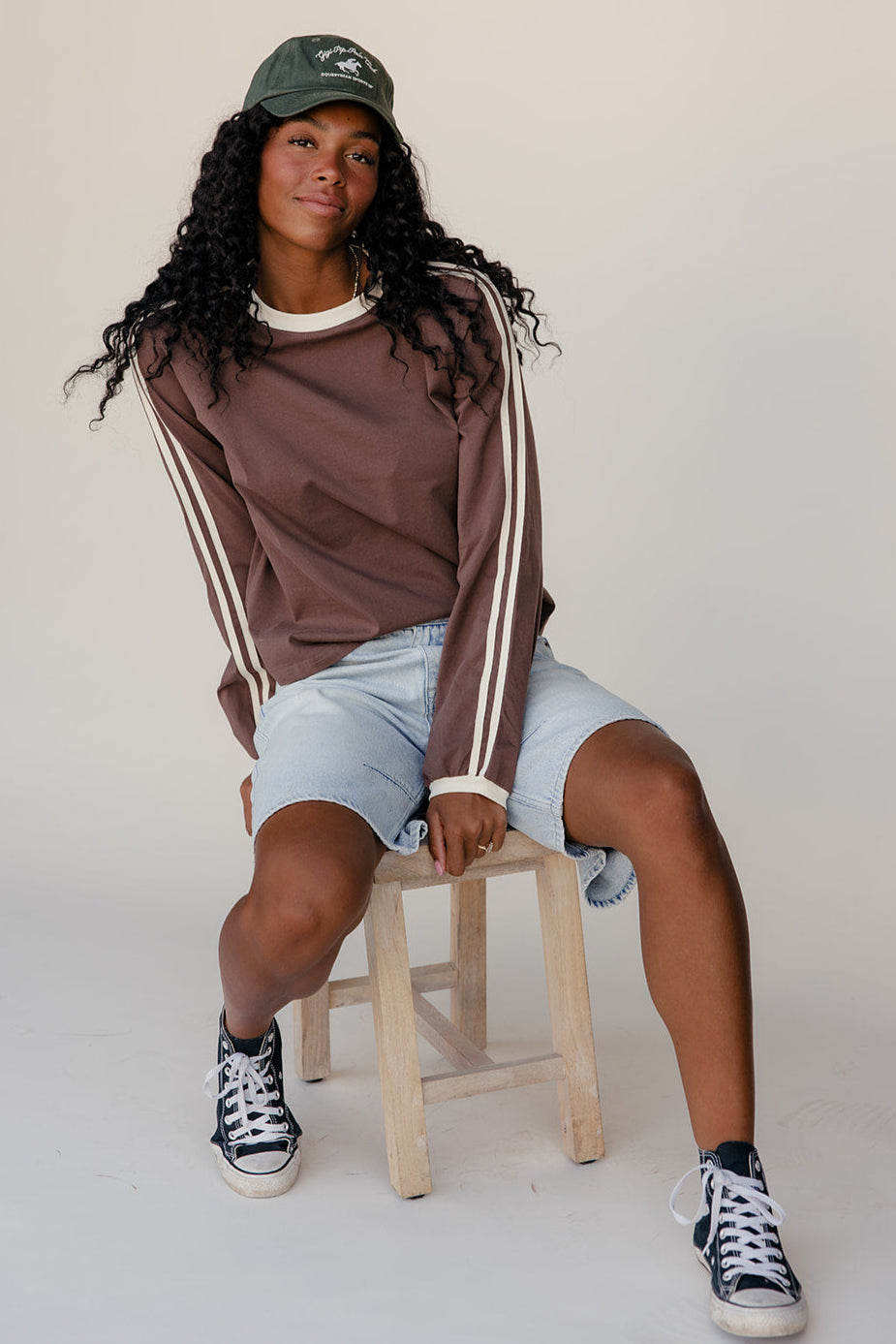 Person wearing a brown sweatshirt with white stripes, light blue shorts, and black sneakers, sitting on a stool against a plain background.