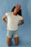 Woman wearing a white t-shirt with orange trim and denim shorts, posing against a blue background.