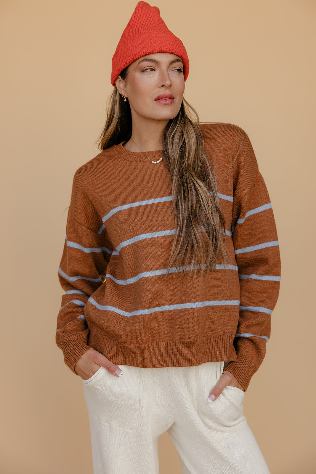 Woman wearing a brown sweater with white stripes and a red beanie against a beige background