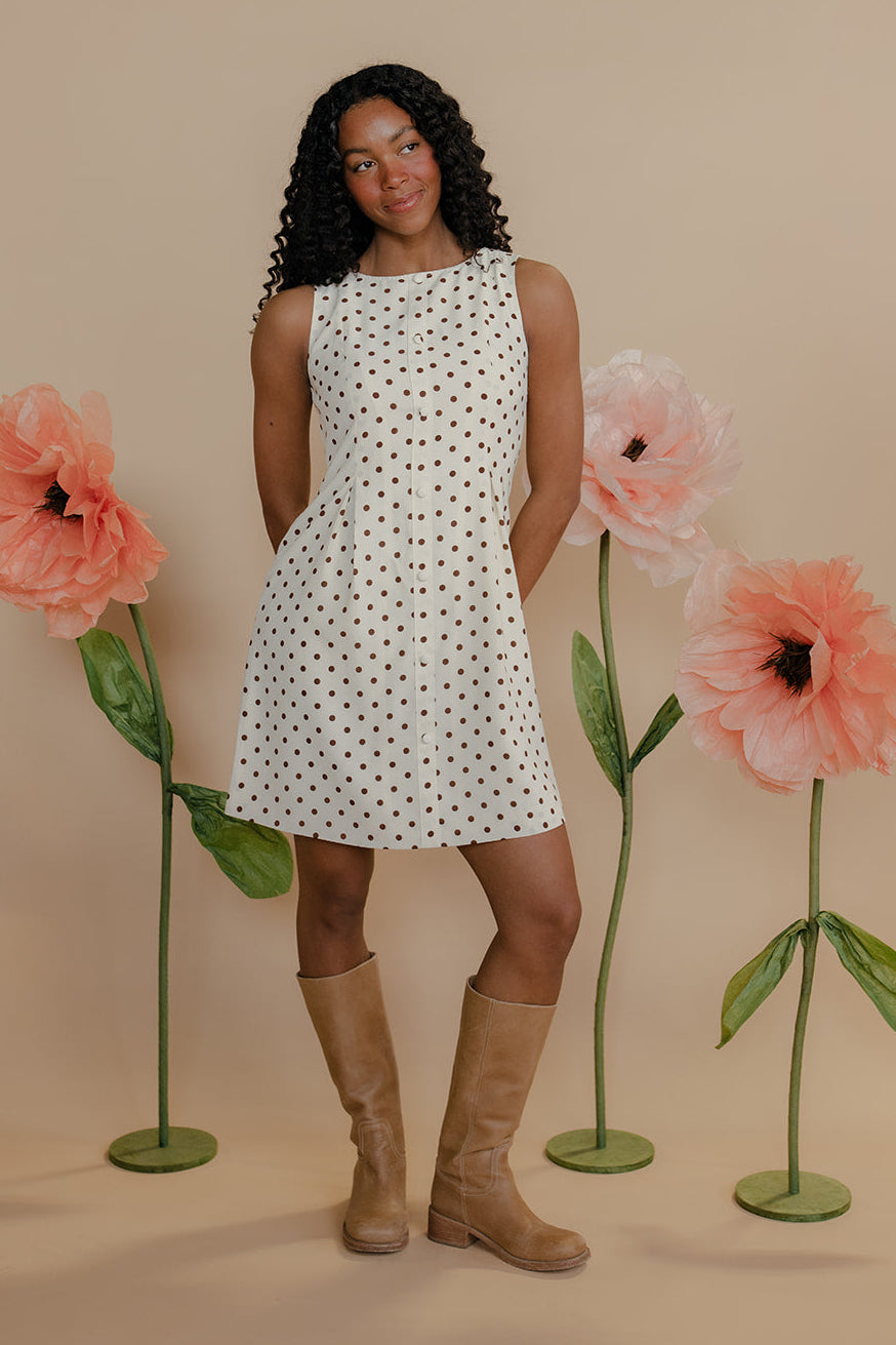 Woman in a polka dot dress standing between large flowers on a beige background