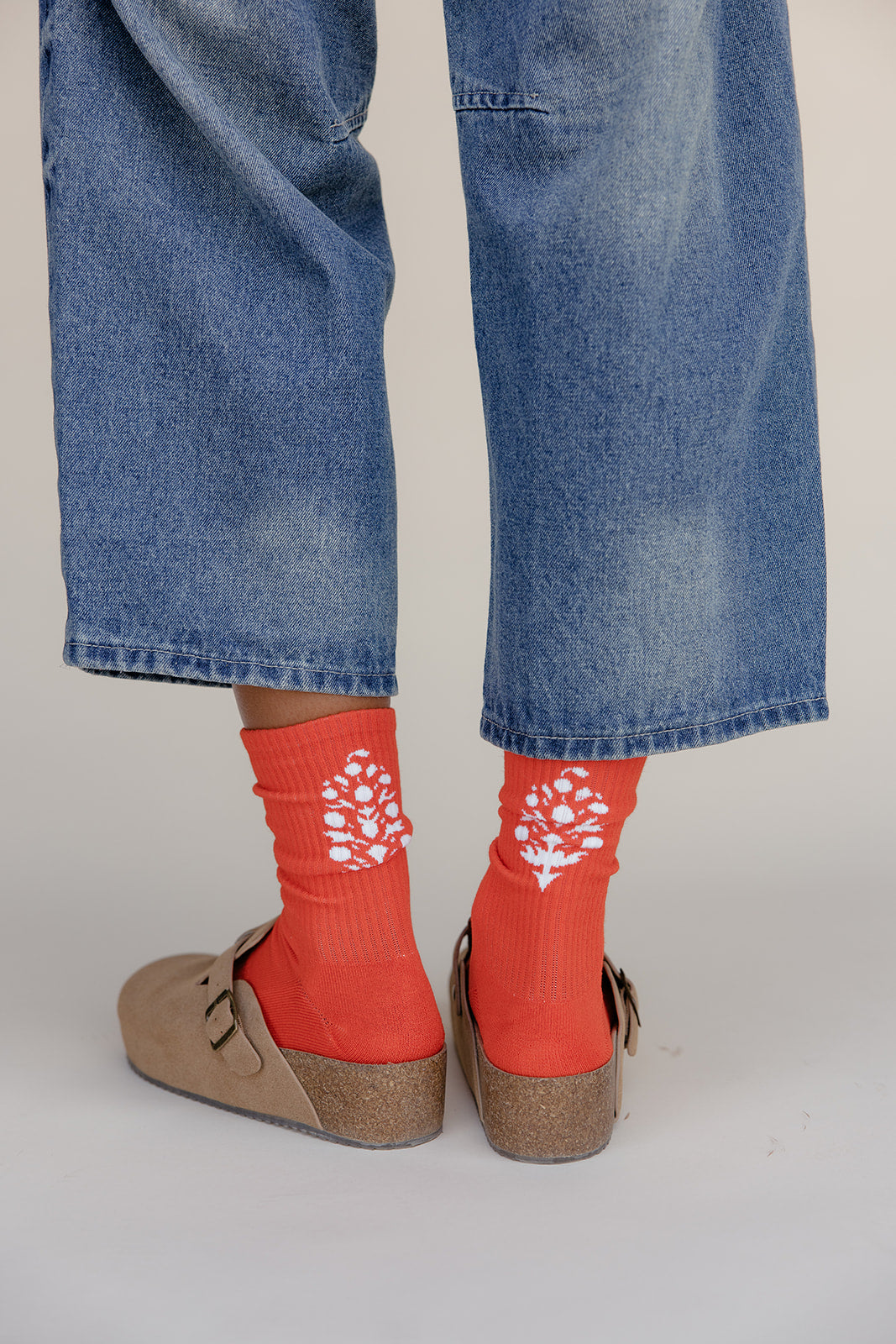 Red socks with white floral patterns worn with blue jeans and brown shoes on a light gray background